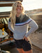 A woman smiling, wearing a striped sweater with a crew neckline, where the stripes feature shades of gray, blue, and purple, and the brand logo 'OK' is visible on the chest.