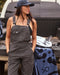 A woman wearing a gray overalls and a dark cap, standing near a vehicle with equipment around her, possibly on a camping trip.