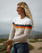 A woman smiling and wearing a beige crewneck sweater with colorful horizontal stripes on the chest area, paired with blue jeans.