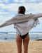 A person standing on a beach wearing a light-colored, oversized shirt with the sleeves extended to the sides and a small black garment in the lower part of the image.