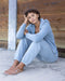 Person wearing a light blue hoodie and pants sitting against a wooden wall.
