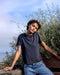 Person wearing a dark gray t-shirt and blue jeans standing outdoors with trees and sky in the background