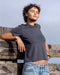 Person wearing a dark gray t-shirt and blue jeans leaning against a wooden fence with a clear sky background.
